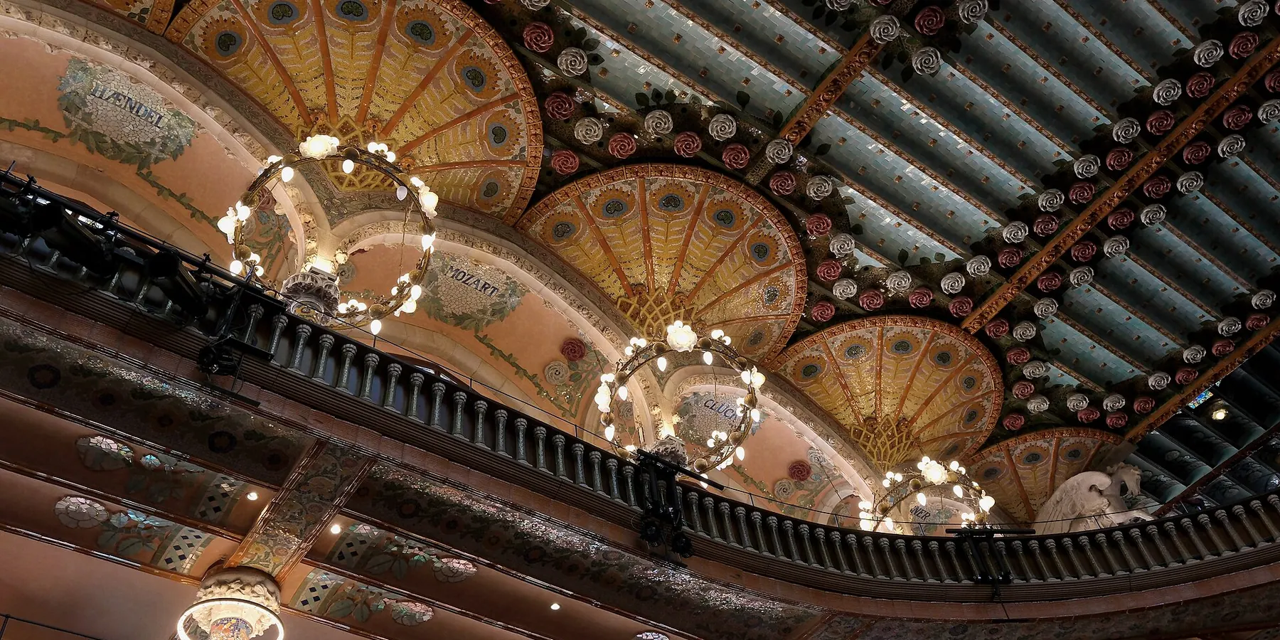 The ceiling of the Palau concert hall with fan-shaped gold and green ornament, ring-shaped hanging lamps and a curved balcony below.