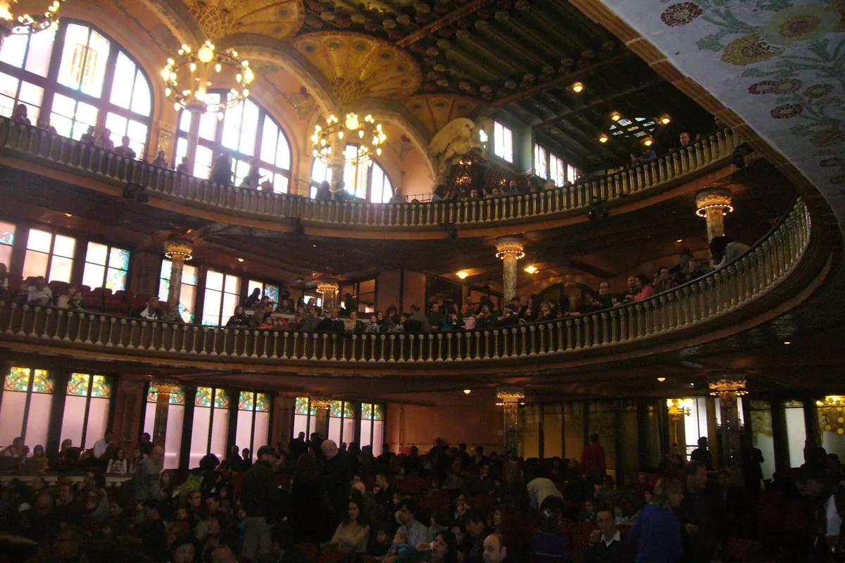 The concert hall of the Palau full of people, with balconies, hanging lamps and arched windows