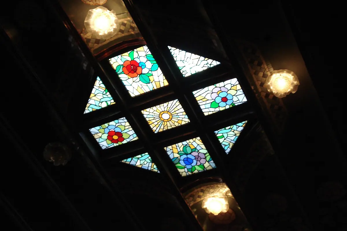 A small stained-glass panel in the Palau ceiling with red, blue and gold flowers set in a diamond pattern between two lamps.