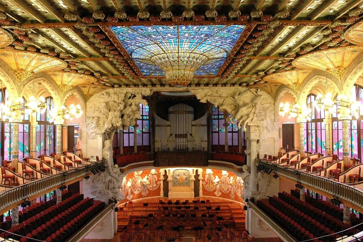 The concert hall of the Palau de la Música Catalana from the stalls, with a stained-glass skylight in blue and gold above and the stage with its muses ahead.
